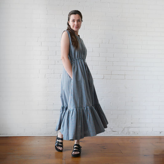 Woman wearing a blue checkered dress standing against a white brick wall.