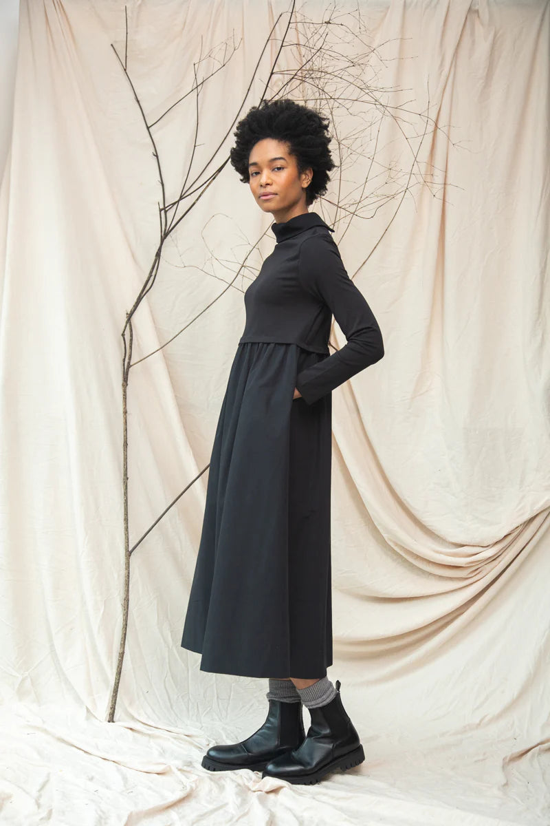 Woman wearing a long black dress standing against a beige draped curtain.