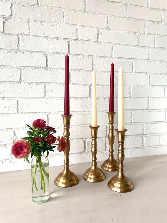 Set of four brass candlesticks with candles against a white brick wall.