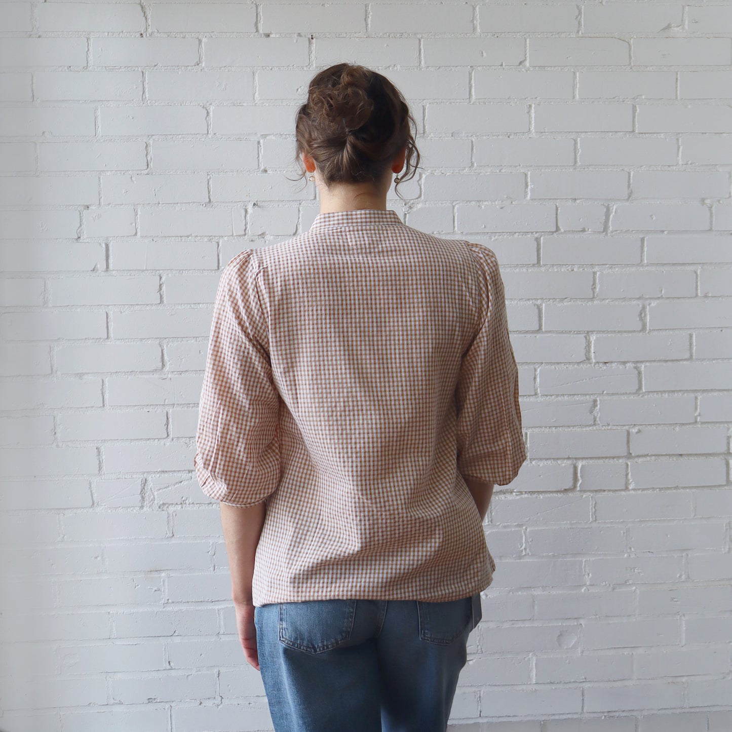 Person wearing a checkered shirt and blue jeans standing against a white brick wall.
