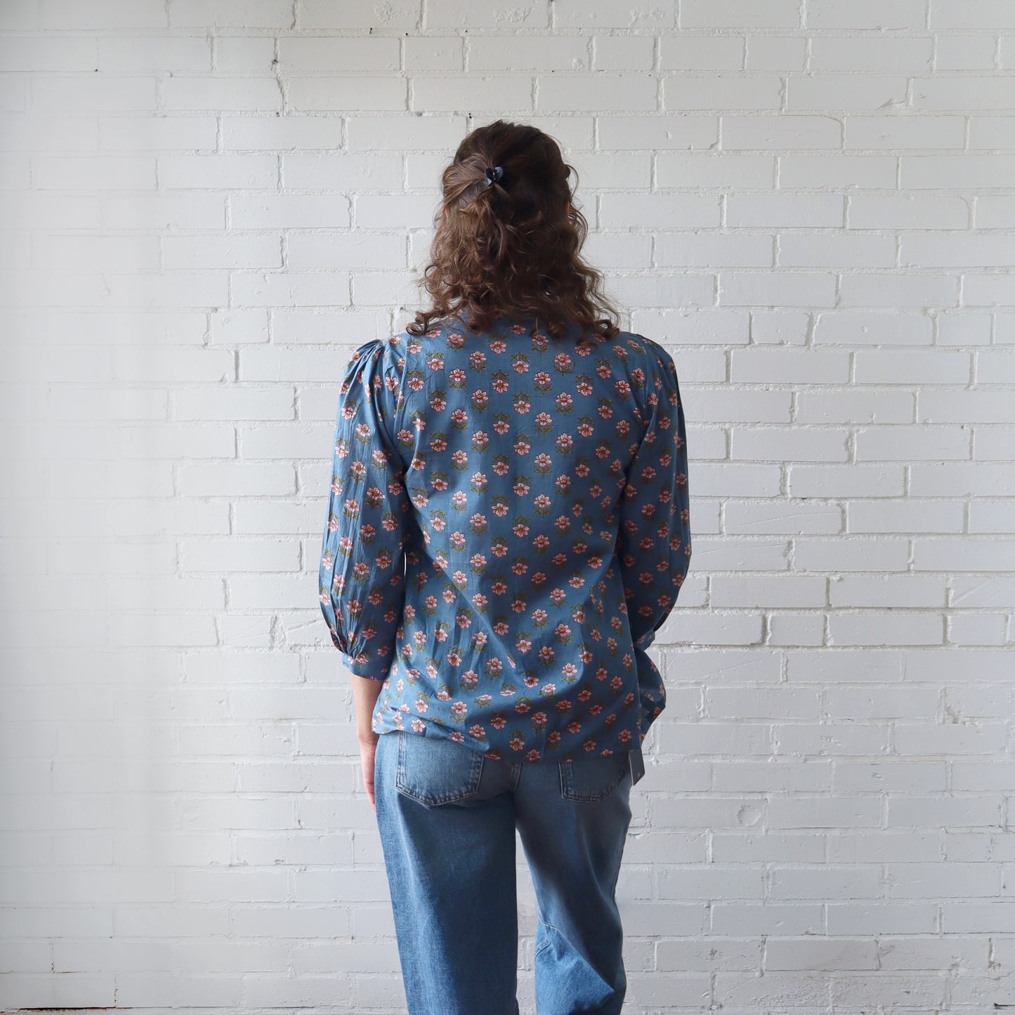 Person wearing a blue patterned blouse and jeans standing against a white brick wall.