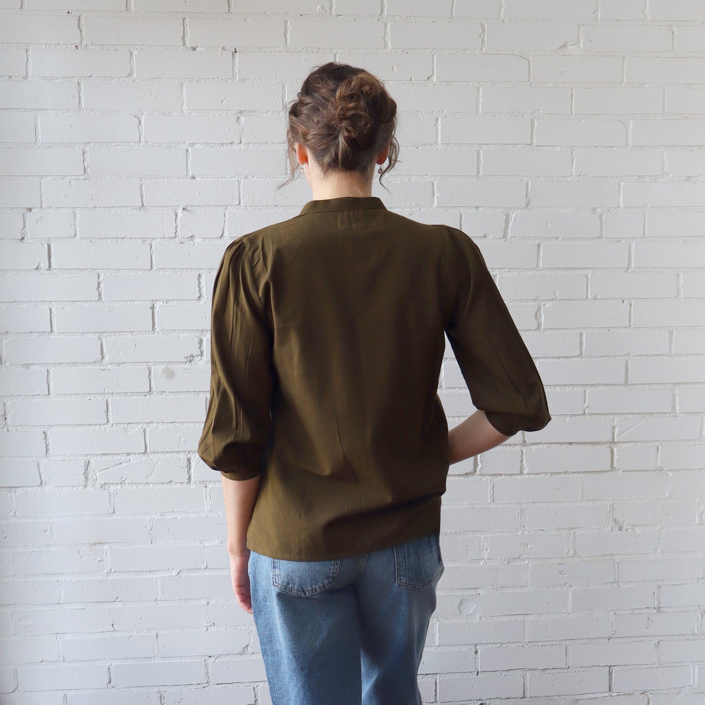 Person wearing a brown shirt and blue jeans against a white brick wall