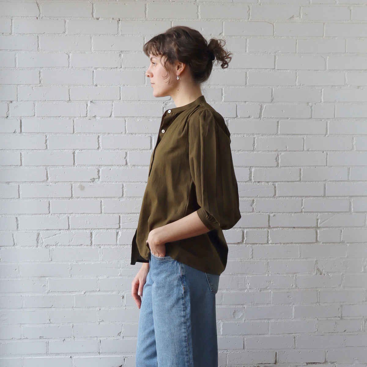 Person wearing an olive green shirt and blue jeans against a white brick wall.
