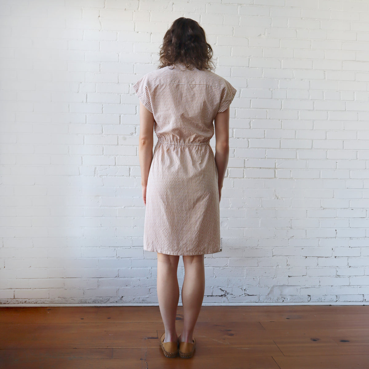 Woman wearing a beige checkered dress standing against a white brick wall.