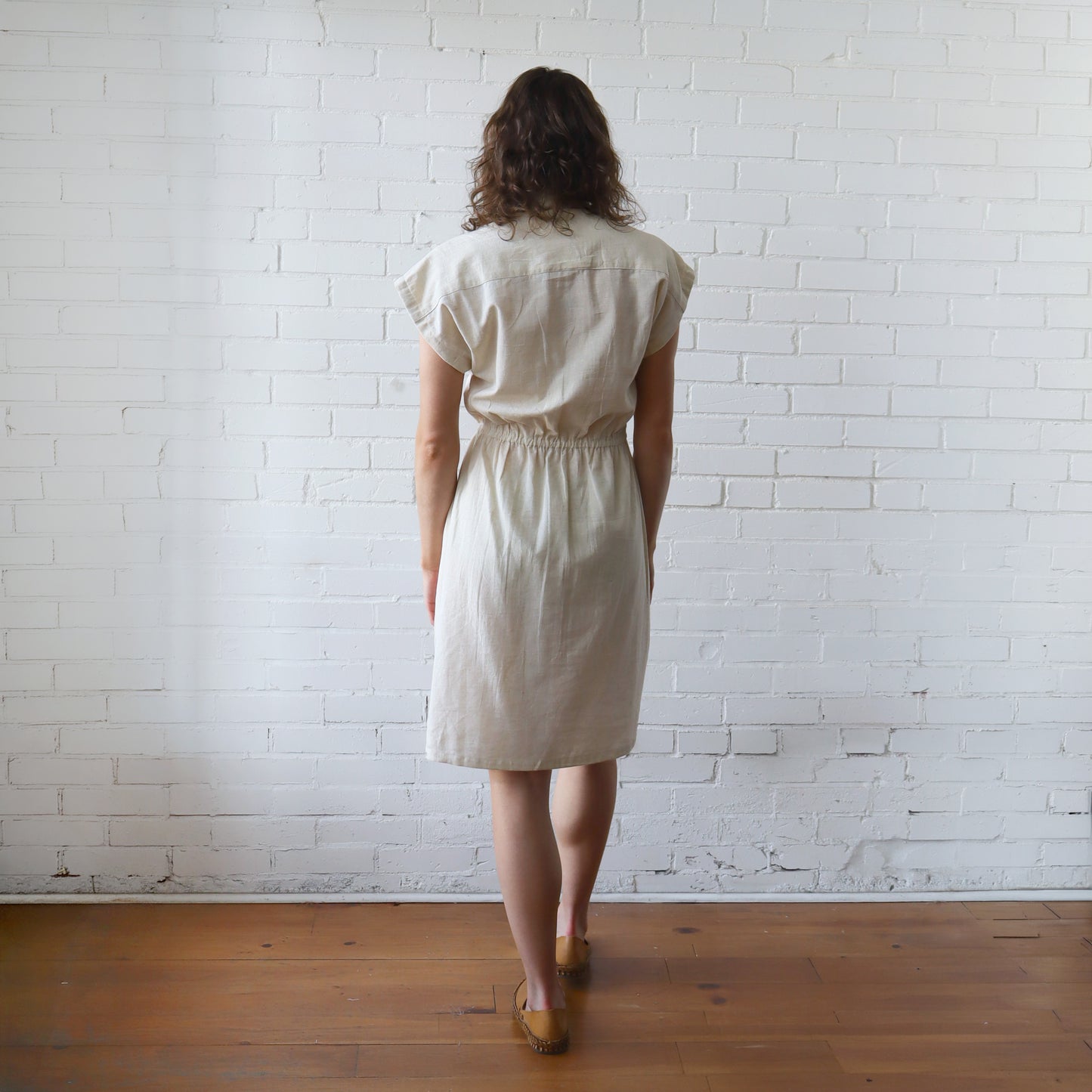 Woman wearing a beige dress standing against a white brick wall.