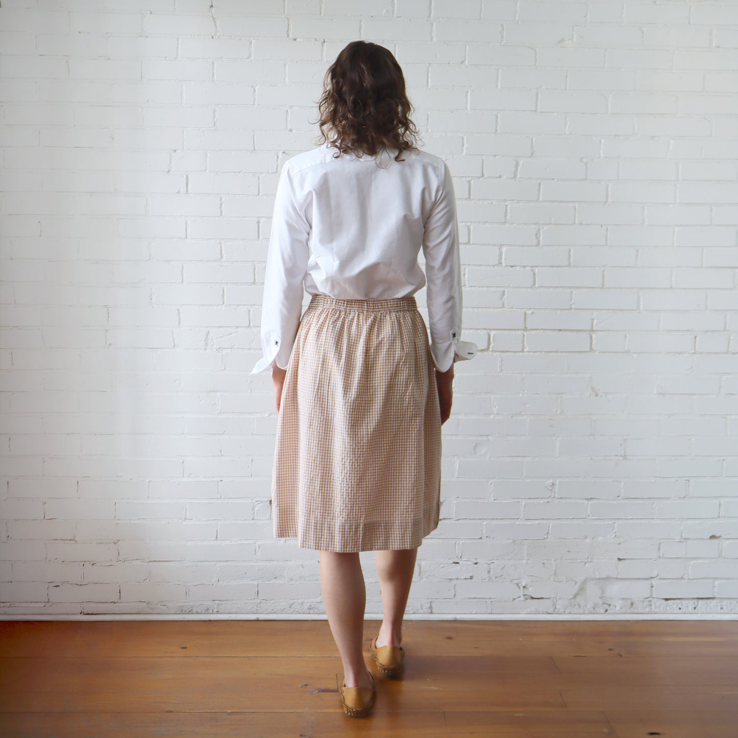 Person wearing a white shirt and beige checkered skirt standing against a white brick wall.