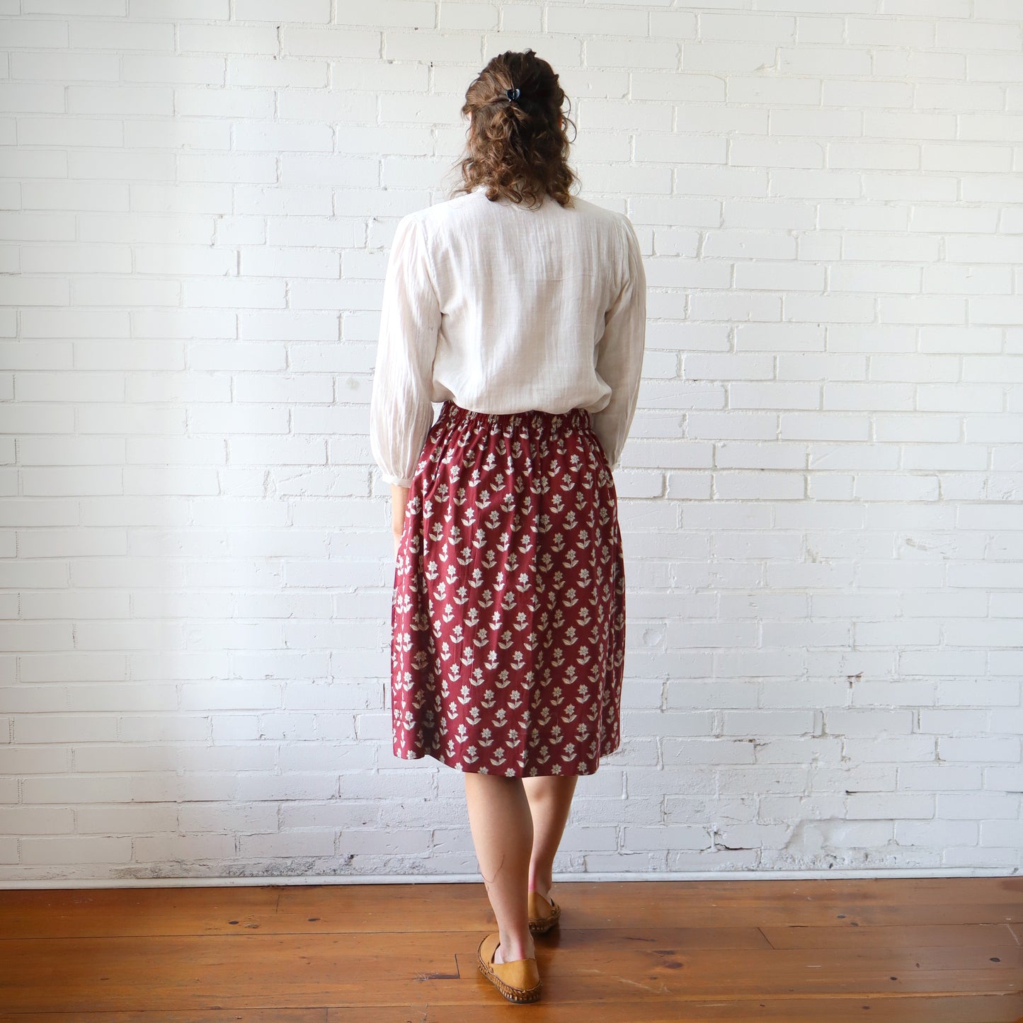 Person wearing a white blouse and red patterned skirt standing against a white brick wall.