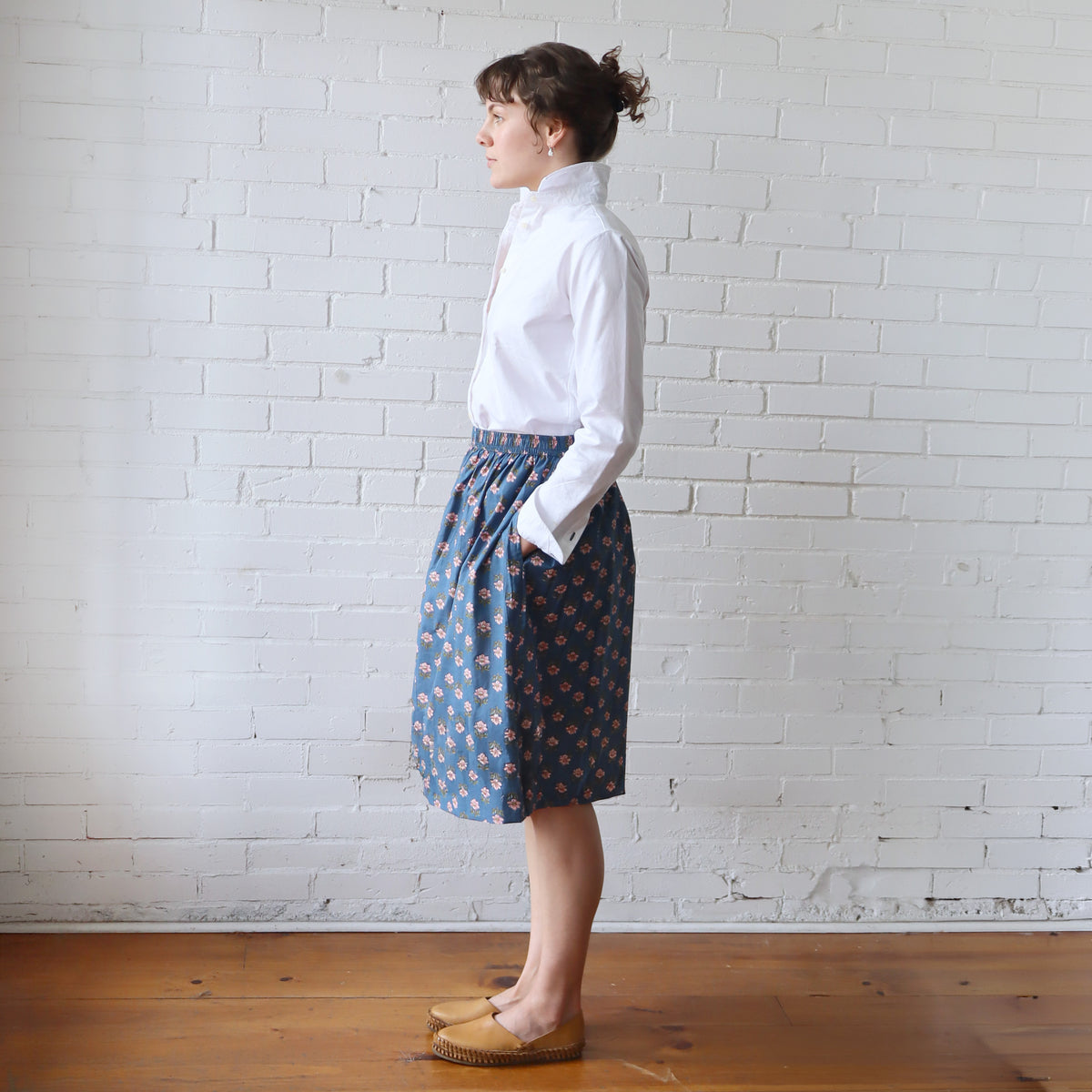 Woman wearing a white blouse and blue patterned skirt against a white brick wall.