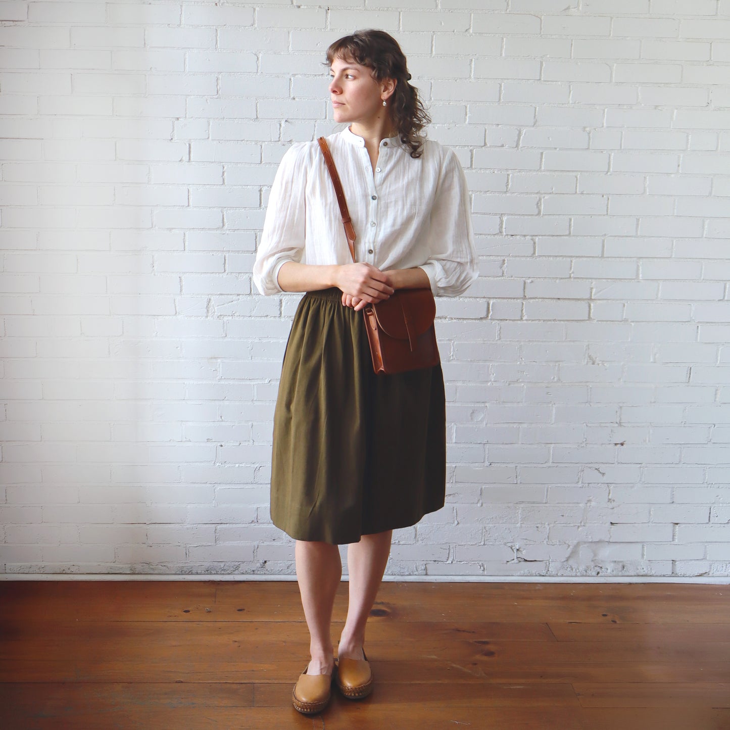 Woman wearing a white shirt and green skirt with a brown bag against a white brick wall.