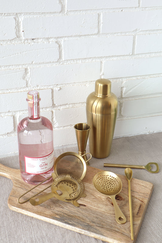 Gold cocktail shaker and tools on a wooden board with a bottle of pink gin against a white brick wall.