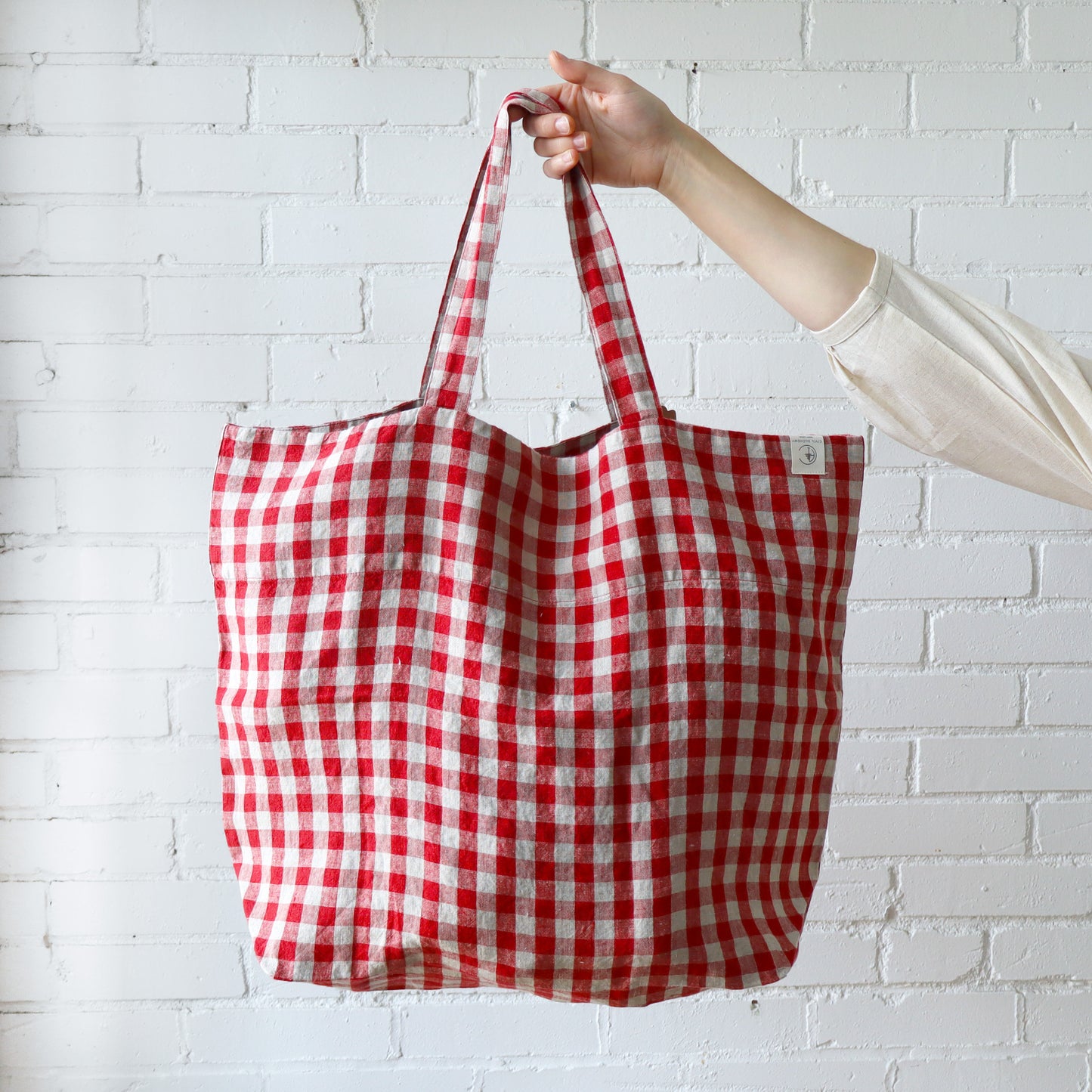 Red and white checkered tote bag held by a person against a white brick wall.