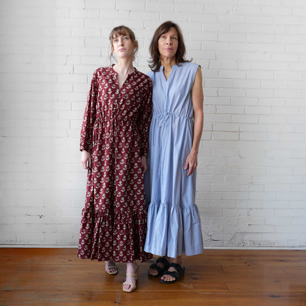 Two women wearing long dresses standing against a white brick wall.