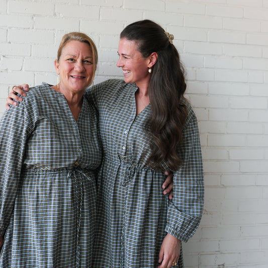 Two women wearing matching plaid dresses standing against a white brick wall.