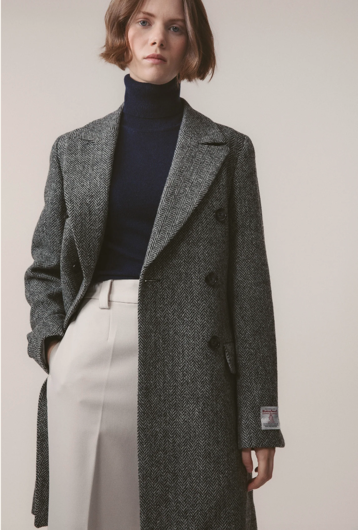 Person wearing a gray textured coat over a navy turtleneck and white pants on a plain background