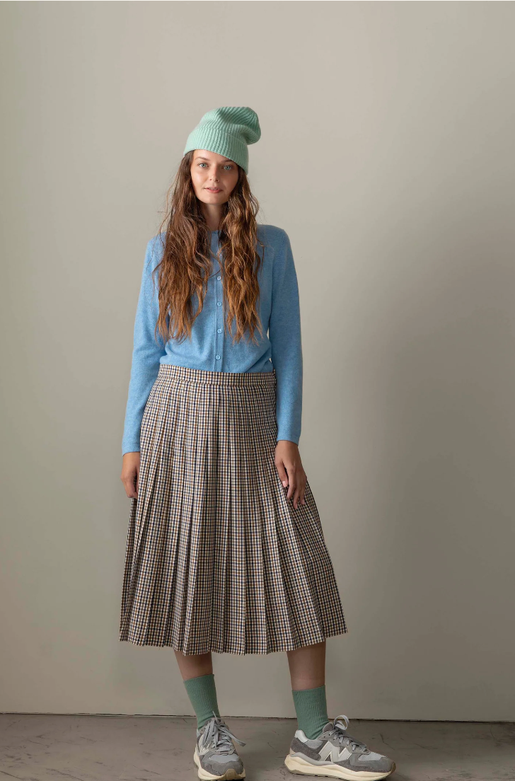 Woman wearing a blue sweater, plaid skirt, and green beanie against a plain background
