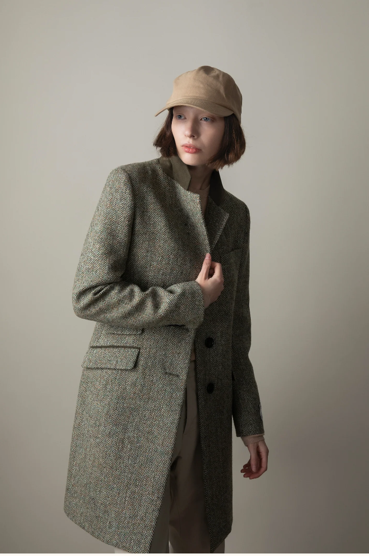 Person wearing a long green coat and beige cap against a plain background