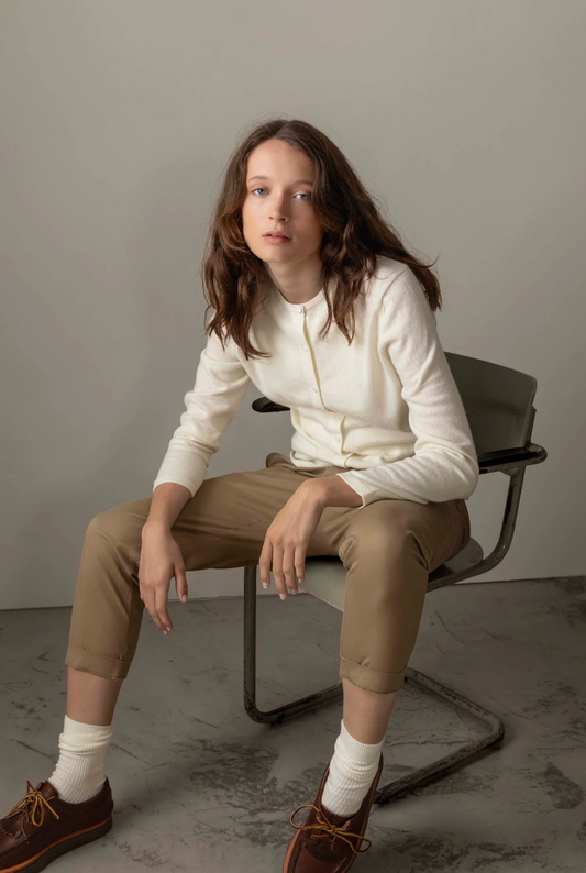 Person sitting on a chair wearing a white sweater and beige pants against a plain background