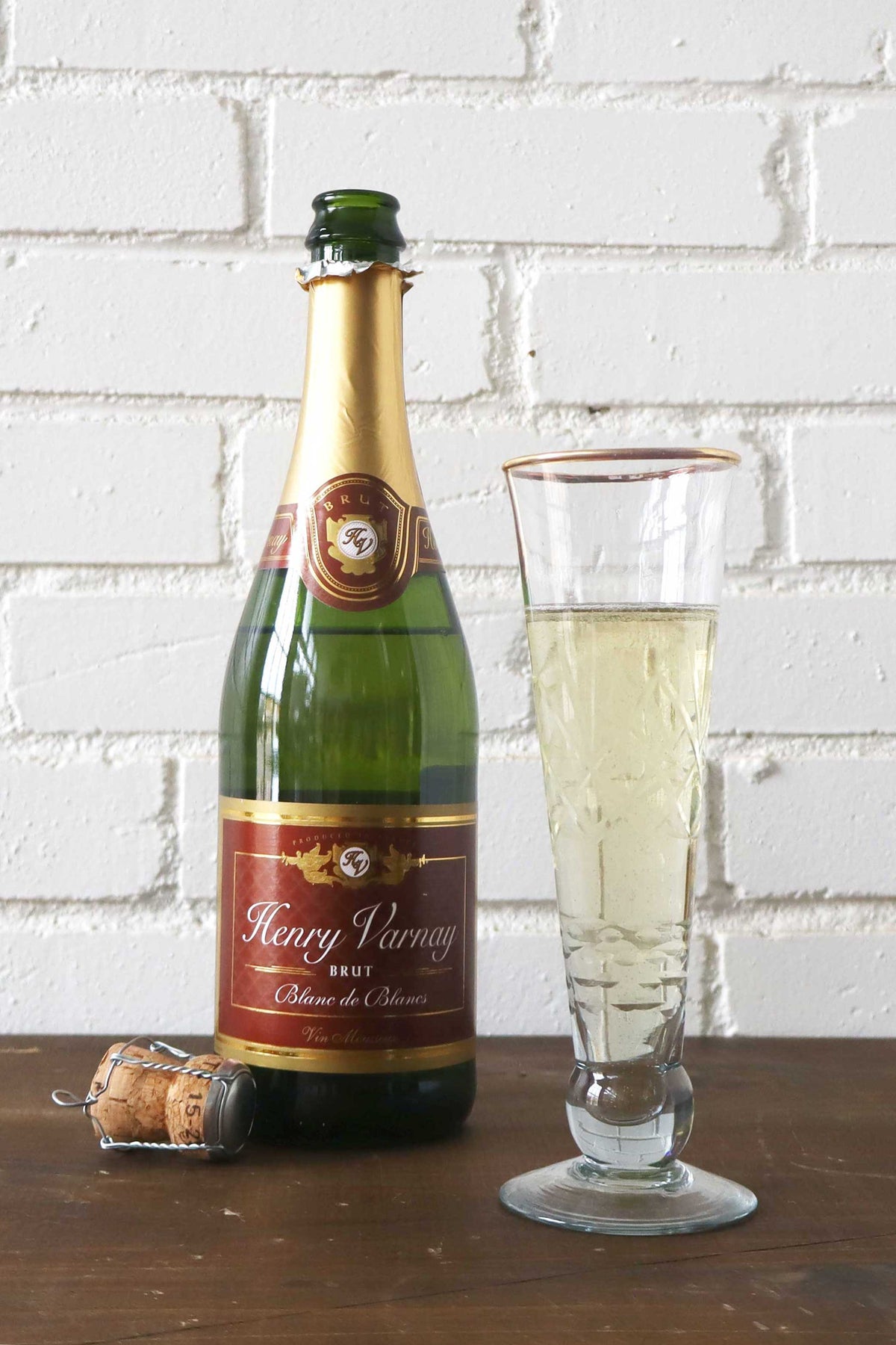 Bottle of Henry Varnay wine with a glass of wine on a wooden surface against a white brick wall.