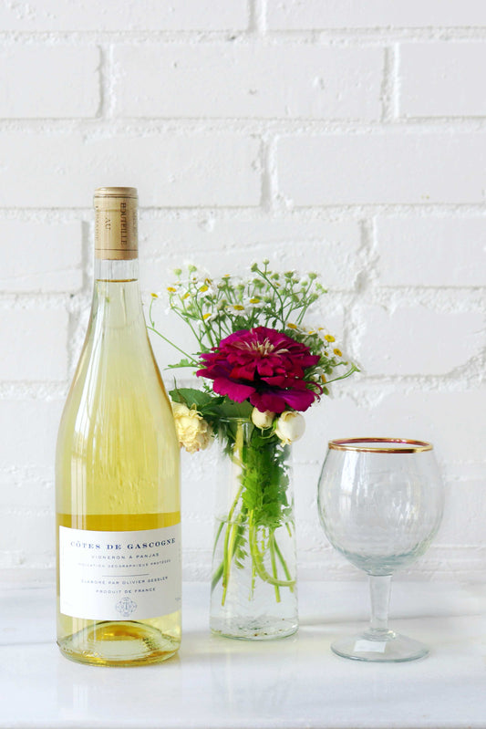 Wine bottle, vase with flowers, and wine glass on a white surface with a white brick wall background