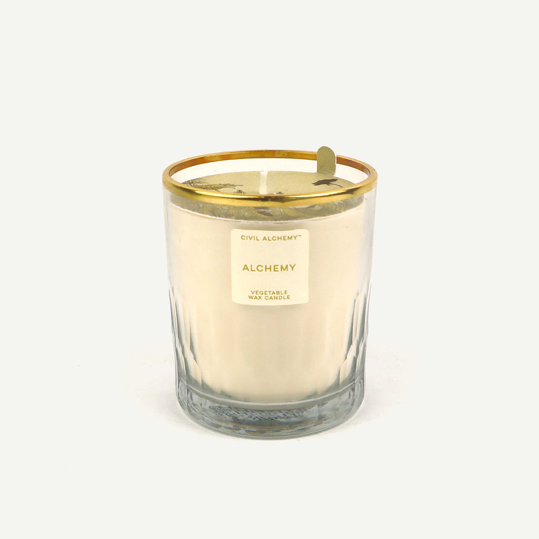 Candle with gold lid and 'Alchemy' label on a white background