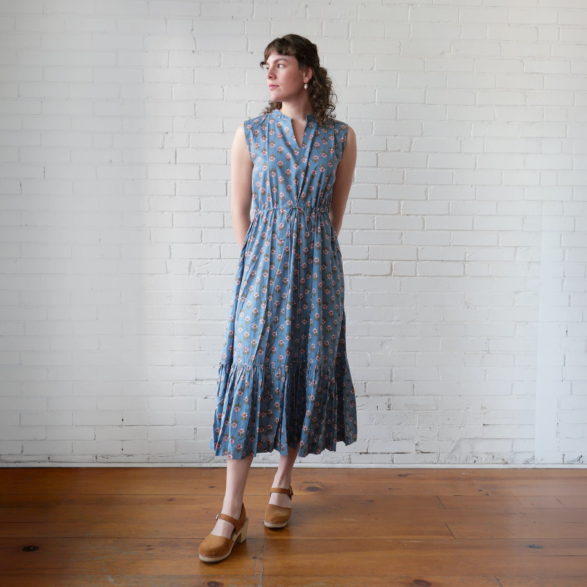 Woman wearing a blue patterned dress standing against a white brick wall.