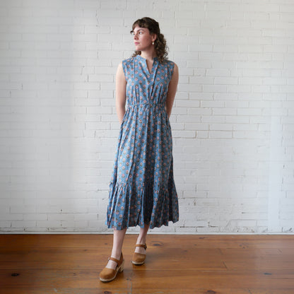 Woman wearing a blue patterned dress standing against a white brick wall.