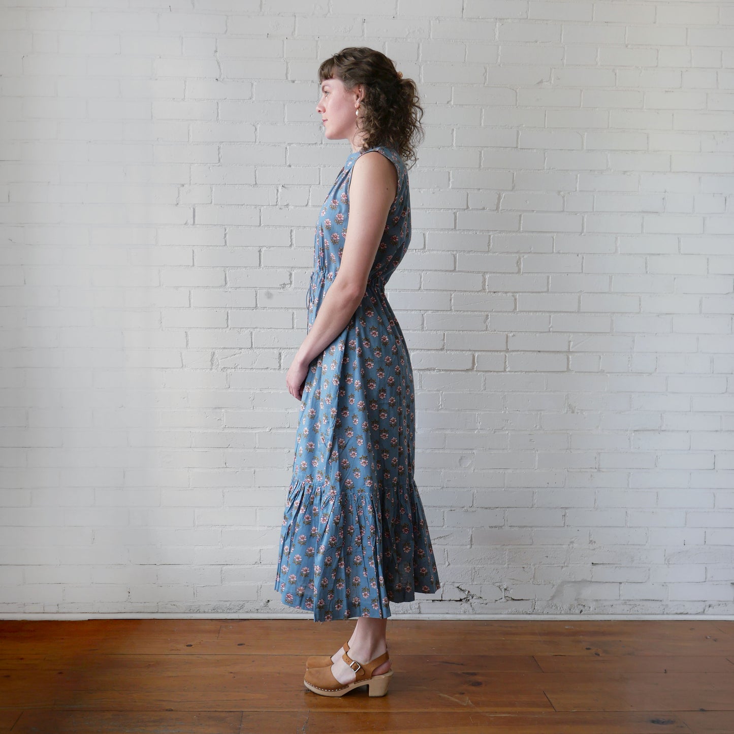 Woman wearing a blue floral dress standing against a white brick wall.