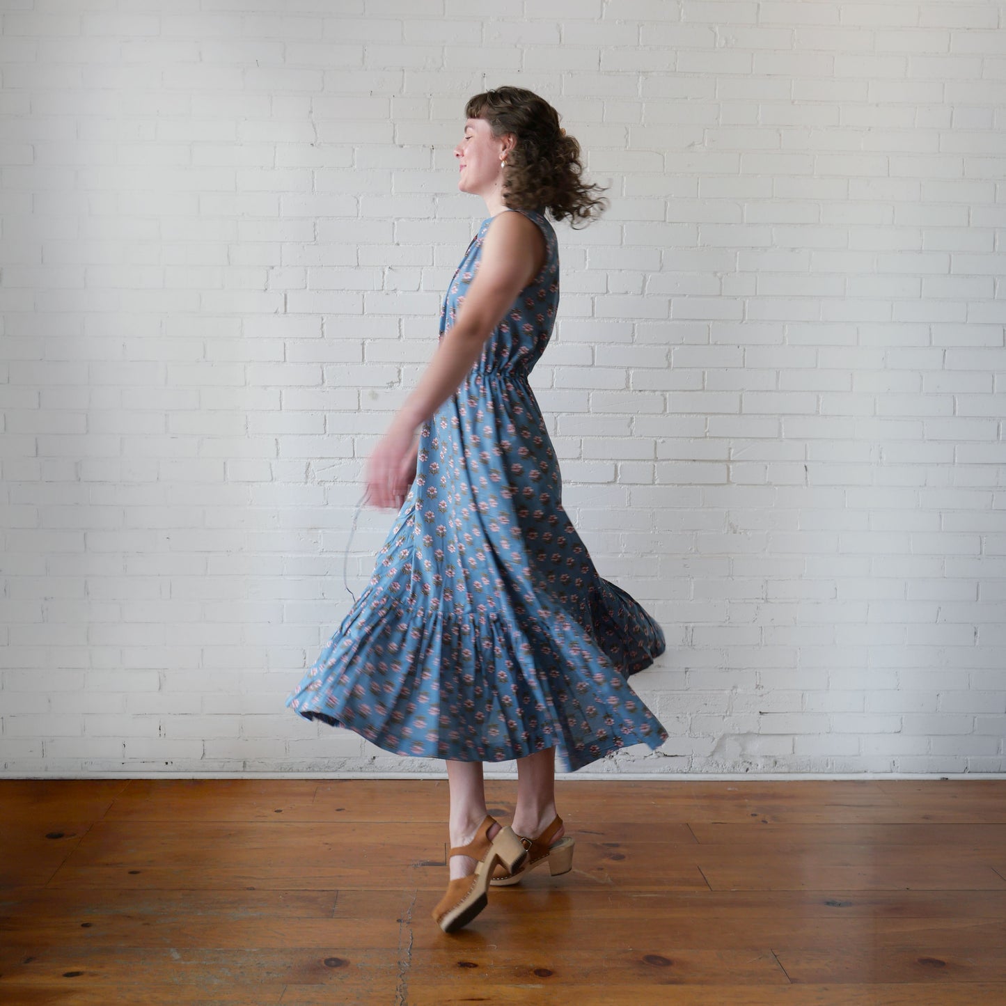 Woman in a blue patterned dress standing against a white brick wall.