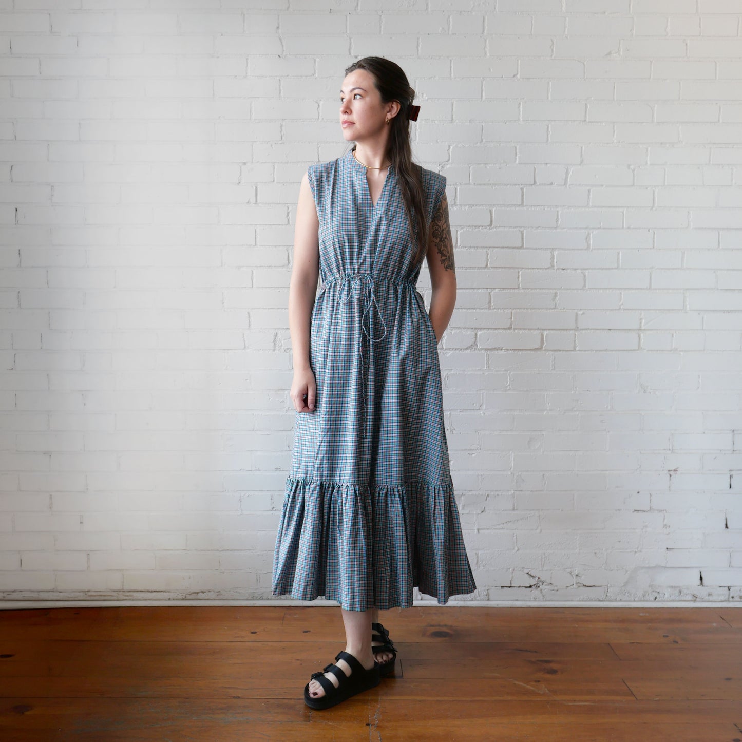 Woman wearing a blue checkered dress standing against a white brick wall.