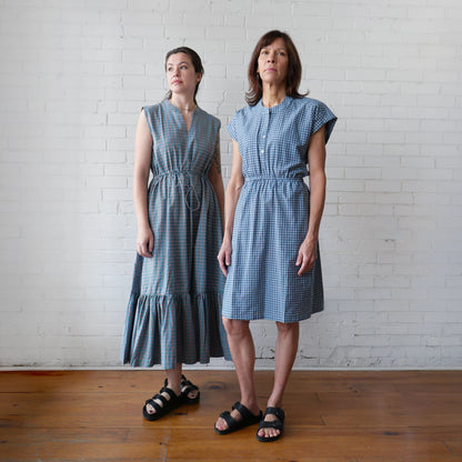 Two women wearing blue checkered dresses standing against a white brick wall.