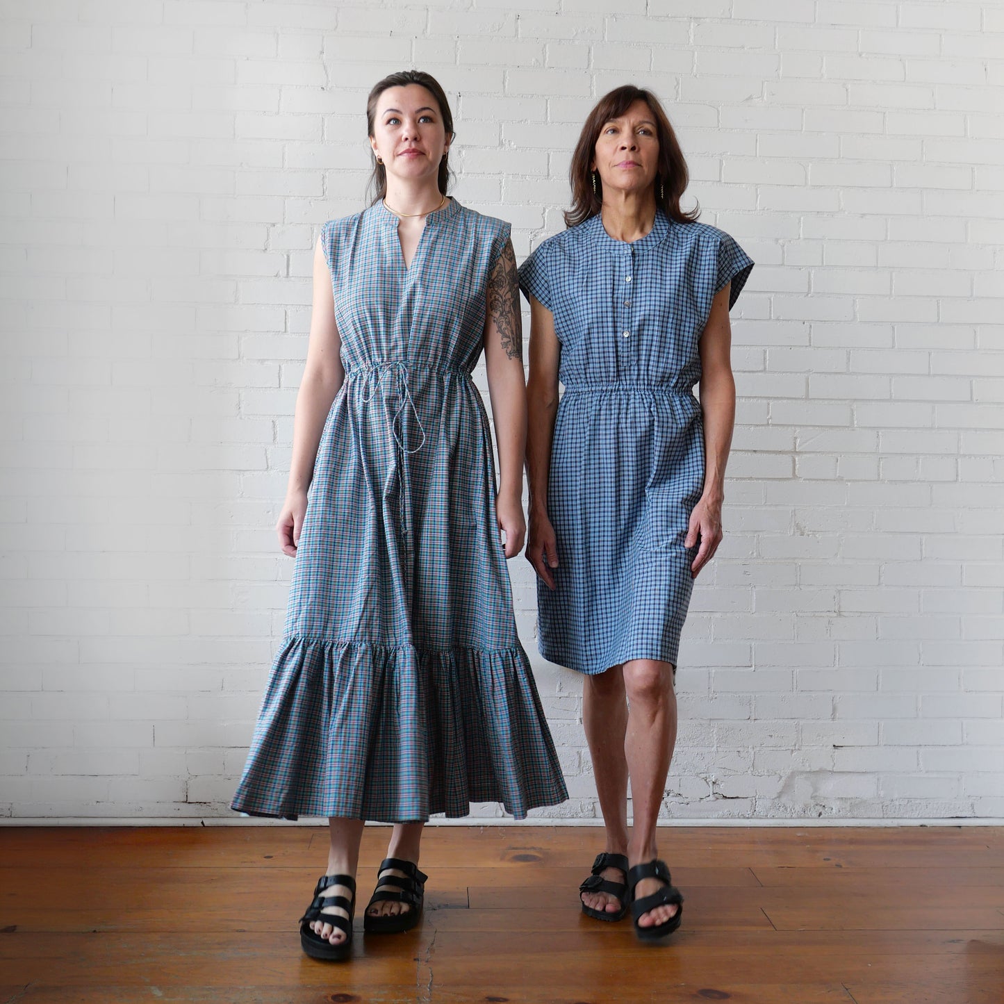 Two women in blue plaid dresses walking against a white brick wall.