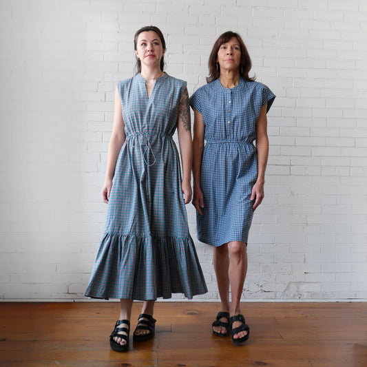 Two women in blue plaid dresses walking against a white brick wall.