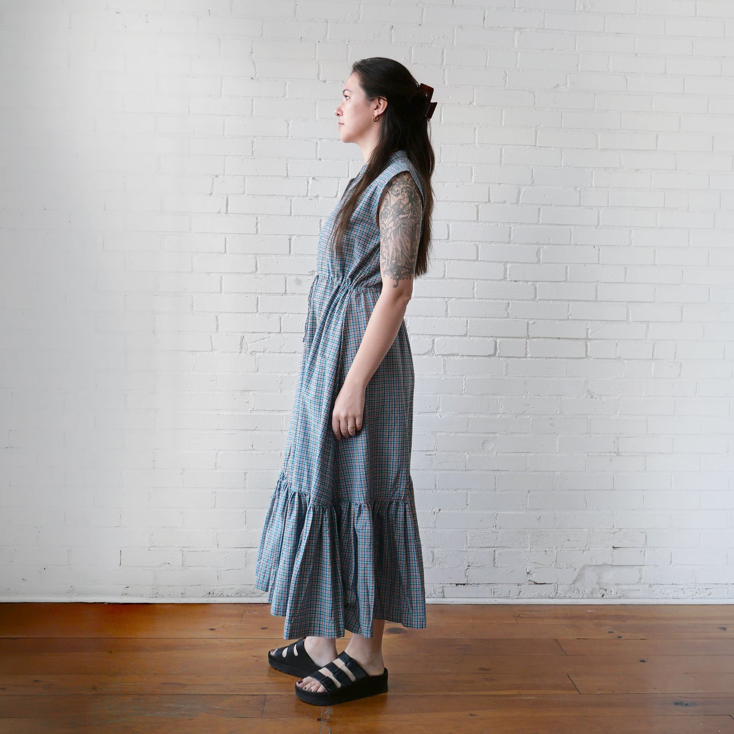 Woman wearing a blue checkered dress standing against a white brick wall.