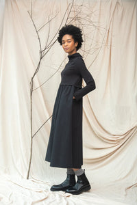 Woman wearing a long black dress standing against a beige draped curtain.
