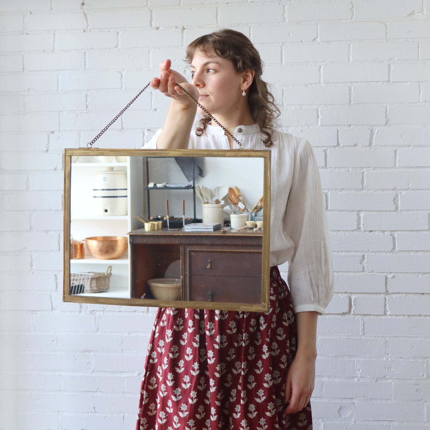 Woman holding a rectangular mirror with a wooden frame against a white brick wall.