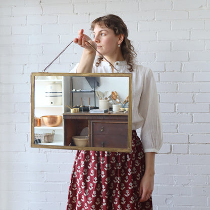 Woman holding a rectangular mirror with a wooden frame against a white brick wall.
