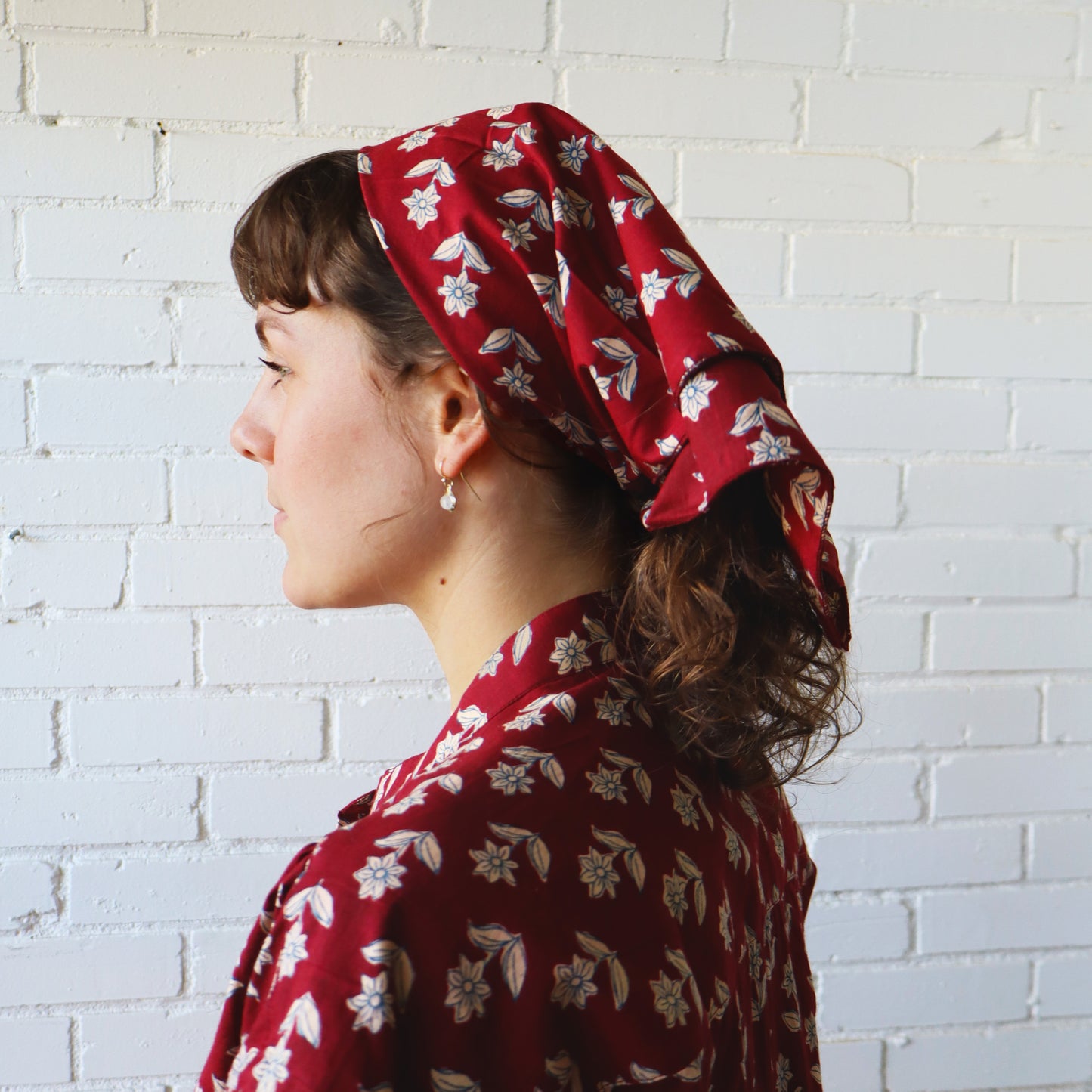 Woman wearing a red floral headscarf and dress against a white brick wall.