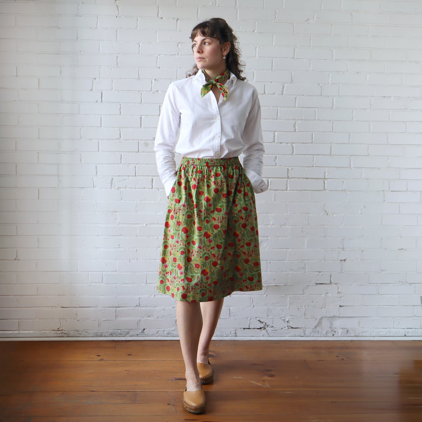 Woman wearing a white shirt and green floral skirt against a white brick wall.