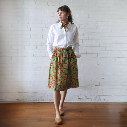 Woman wearing a white shirt and green floral skirt against a white brick wall.