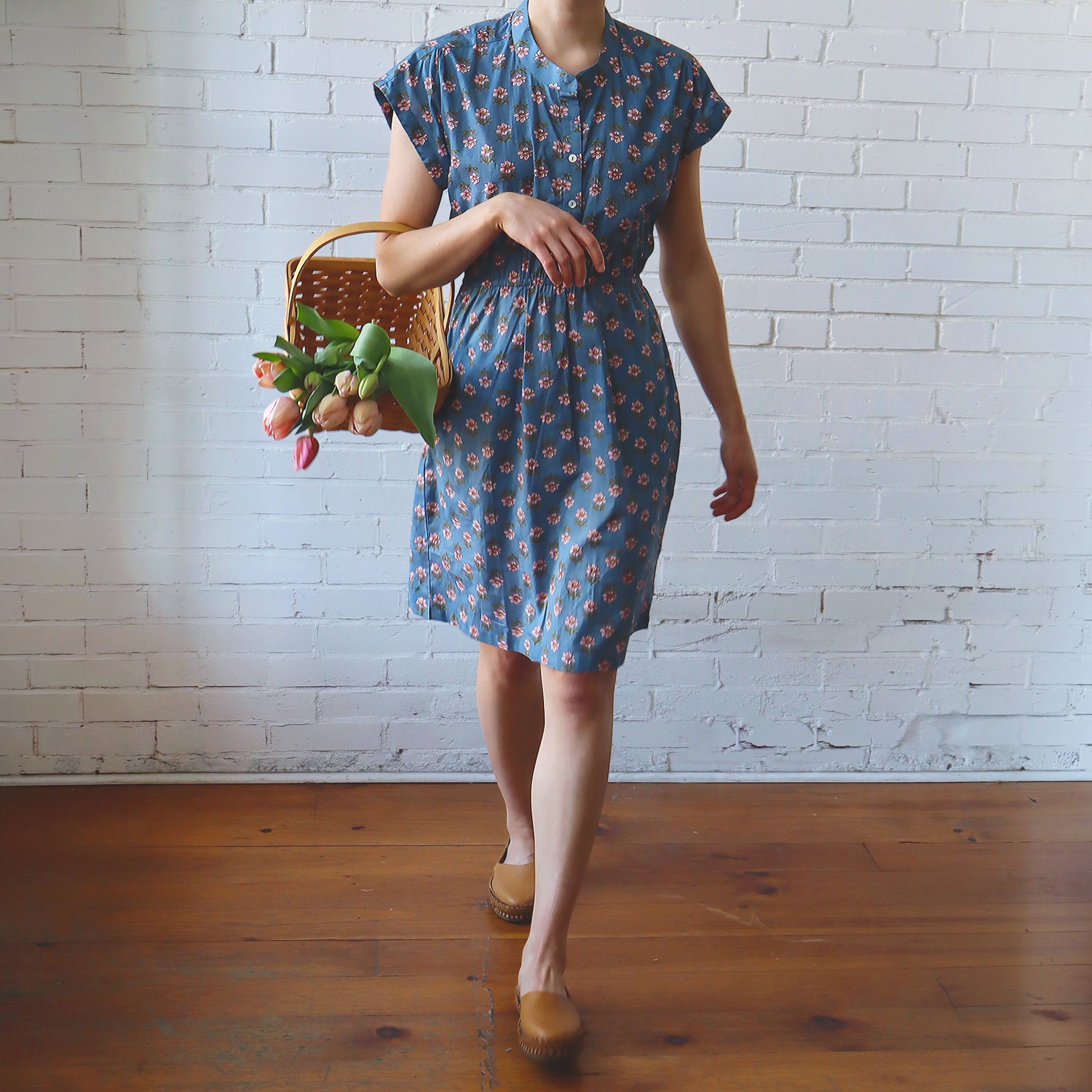 Woman in a blue floral dress holding a basket of flowers against a white brick wall.
