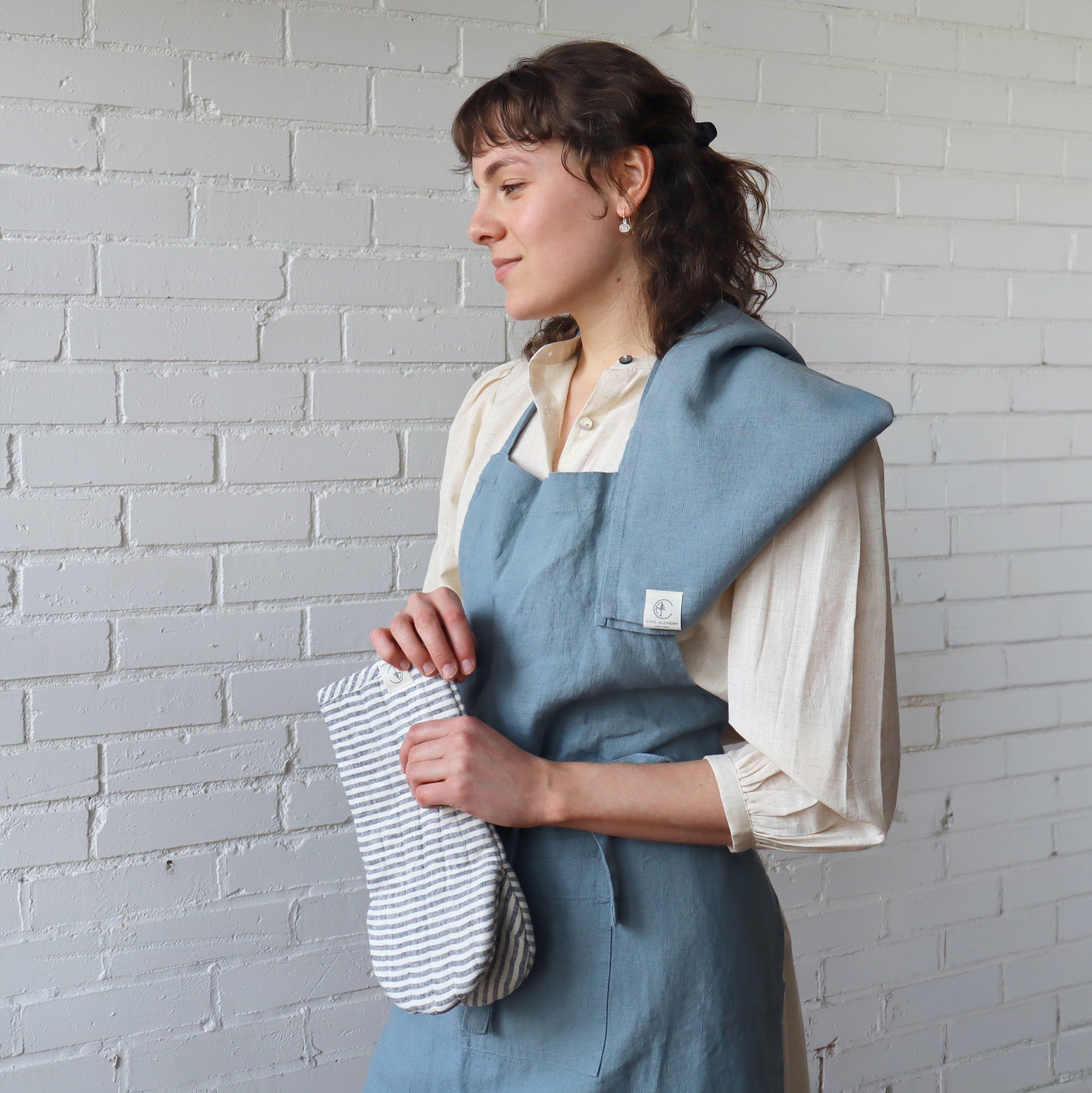Woman wearing a blue apron holding a striped towel against a gray brick wall.