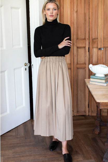 Woman wearing a black turtleneck and Emerson Fry beige pleated skirt in a room with wooden flooring and a wooden door.