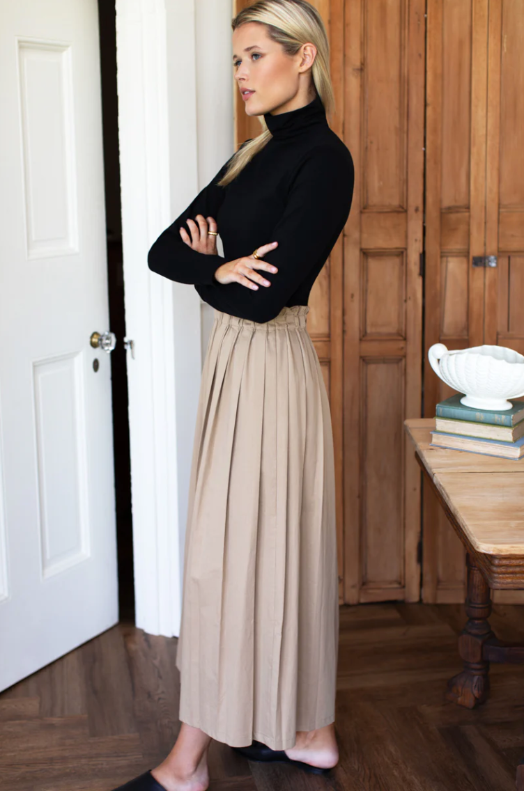 Woman in black top and beige skirt standing in a room with wooden paneling.