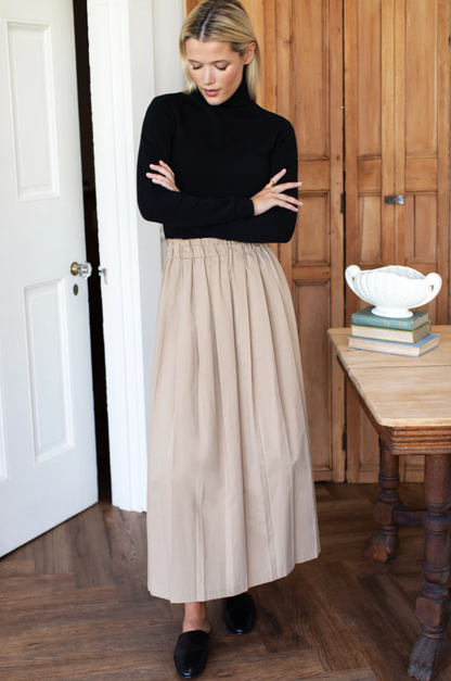 Woman wearing a black turtleneck and beige pleated skirt in a room with wooden flooring and furniture.