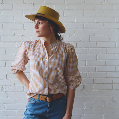 Woman wearing a yellow hat, checkered shirt, and blue jeans against a white brick wall.
