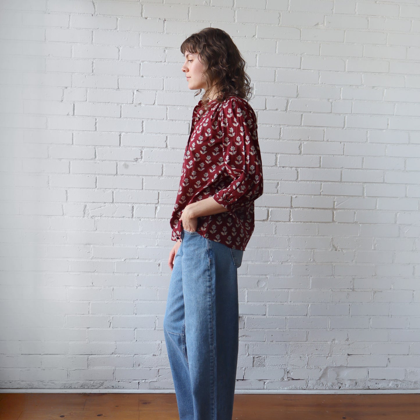 Woman wearing a red patterned blouse and blue jeans against a white brick wall.