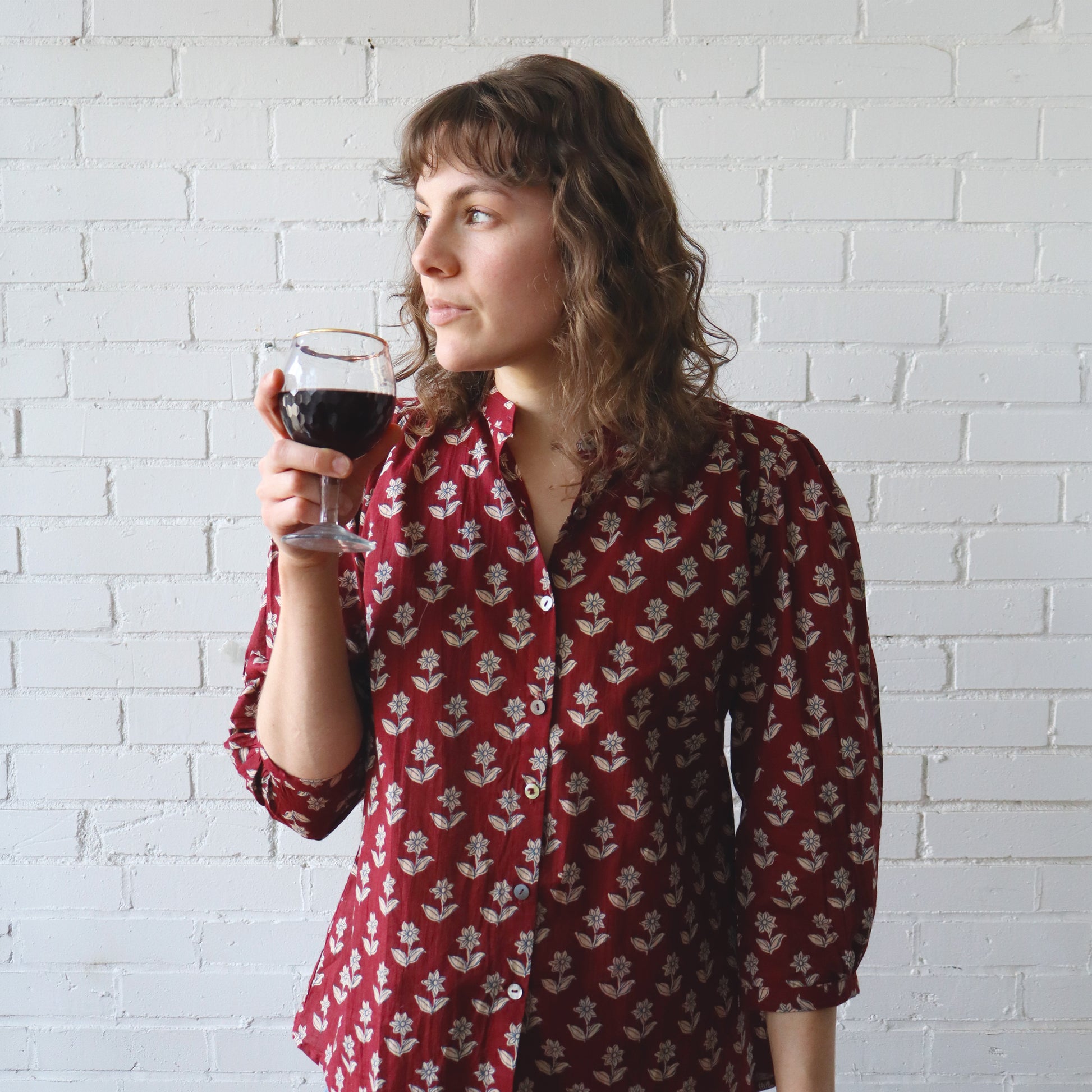 Woman in a red floral blouse holding a glass of red wine against a white brick wall.