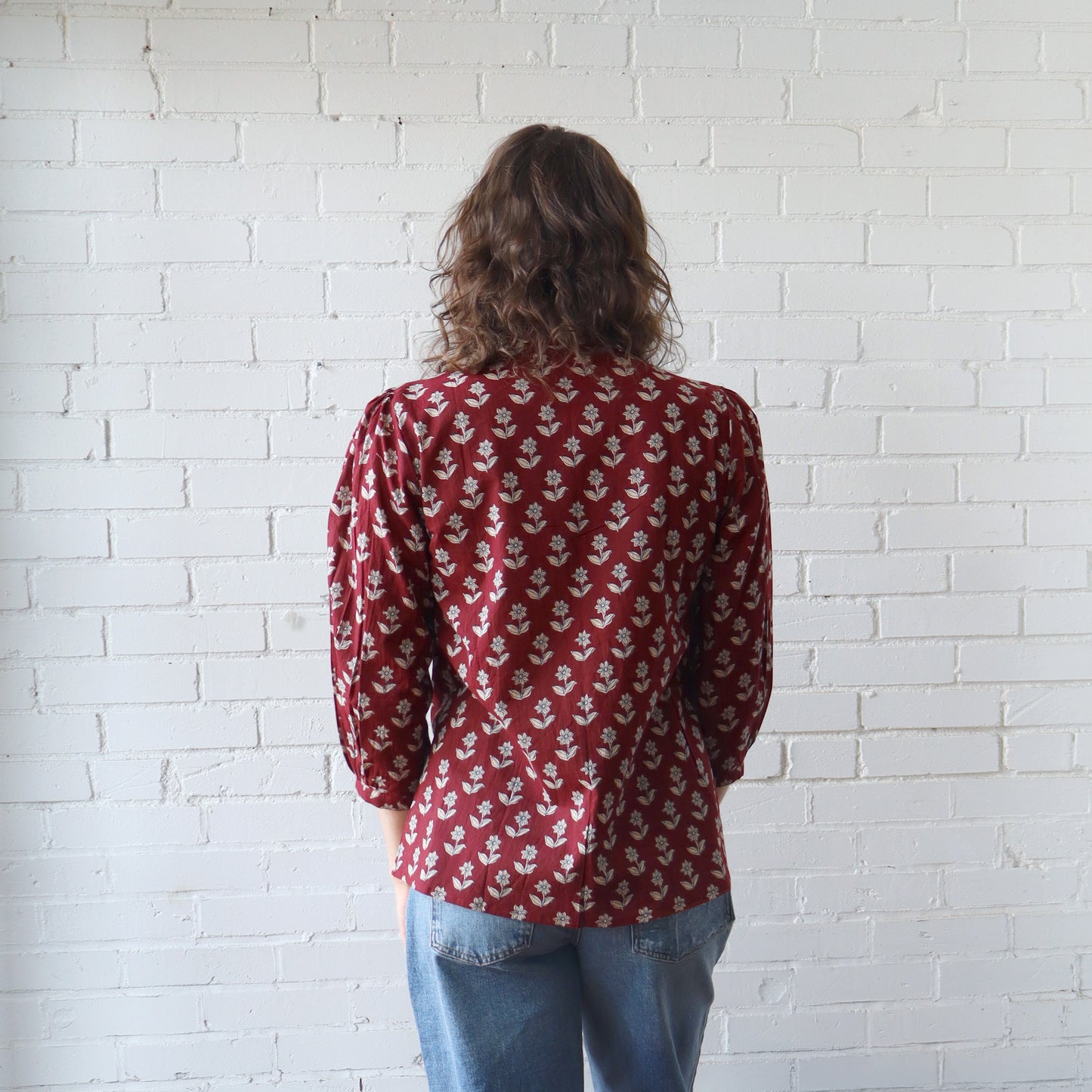 Person wearing a red patterned shirt and blue jeans against a white brick wall.