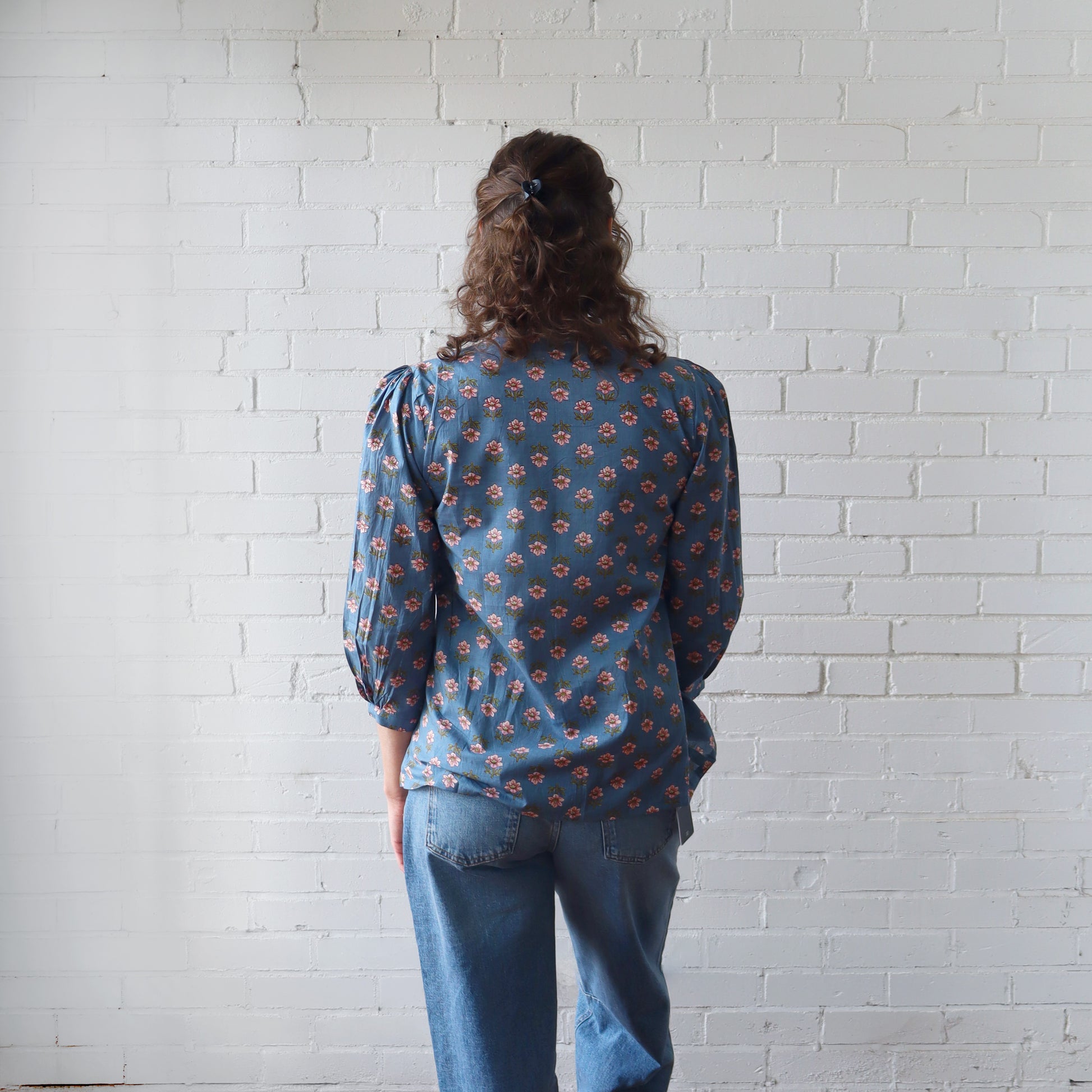 Person wearing a blue patterned blouse and jeans standing against a white brick wall.