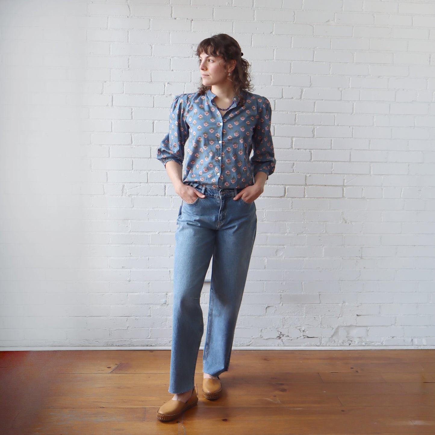 Person wearing a patterned shirt and blue jeans standing against a white brick wall.